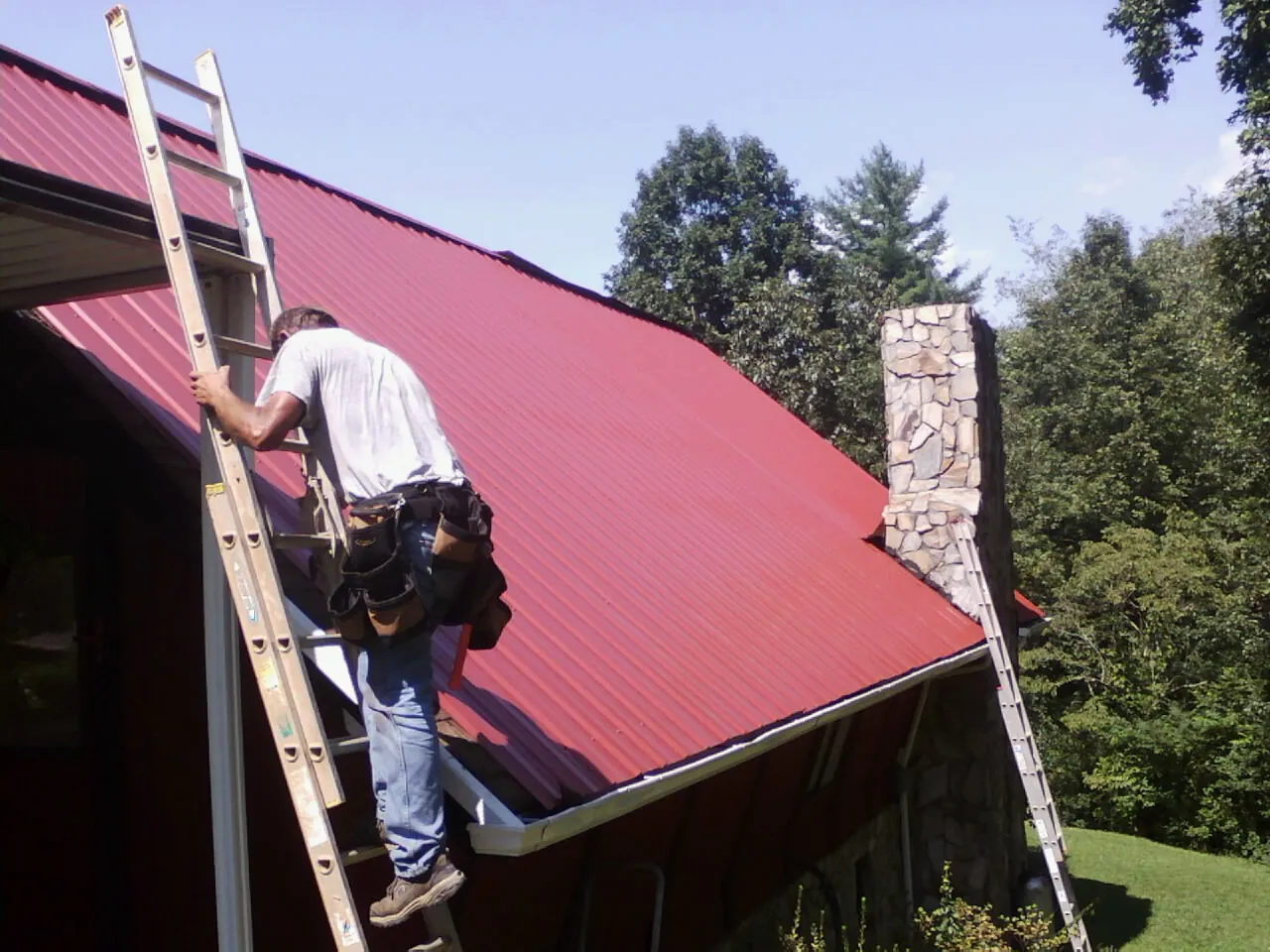 Roofer on a ladder beside a red metal roof and stone chimney — Soffit & Fascia Repair by Roofing Brothers in Mars Hill
